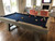 Isaac Slate Pool Table in Silvered Ash showcased in a sunlit open-concept living space with hardwood floors and neutral interiors. The brushed black turnbuckles and tapered legs create a grounded focal point that complements both minimalist and transitional home styles.
