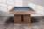 A low, wide-angle lifestyle shot of the Vancouver Slate Pool Table in Honey Ash, drawing the eye to its distinctive reeded wood legs and grounded silhouette. This architectural detail anchors the table with a modern, sculptural presence—perfect for design-savvy shoppers looking for “pool tables with decorative wood base,” “modern reeded leg game tables,” or “high-end slate pool tables with bold furniture styling.”

